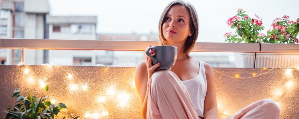 Vrouw zit op haar terras met een kop koffie en lampjes achter haar
