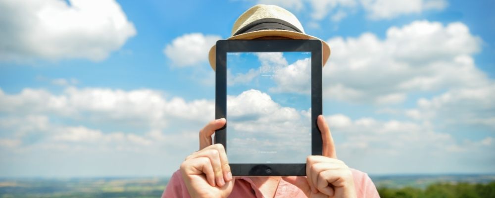Man die een tablet voor zijn hoofd houdt waarop wolken staan waardoor het lijkt alsof hij geen hoofd heeft omdat de achtergrond dezelfde wolken zijn