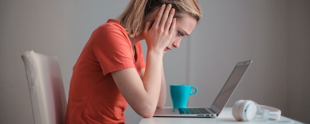 Vrouw die met haar handen tegen haar slaap achter haar laptop zit en naar het scherm staart
