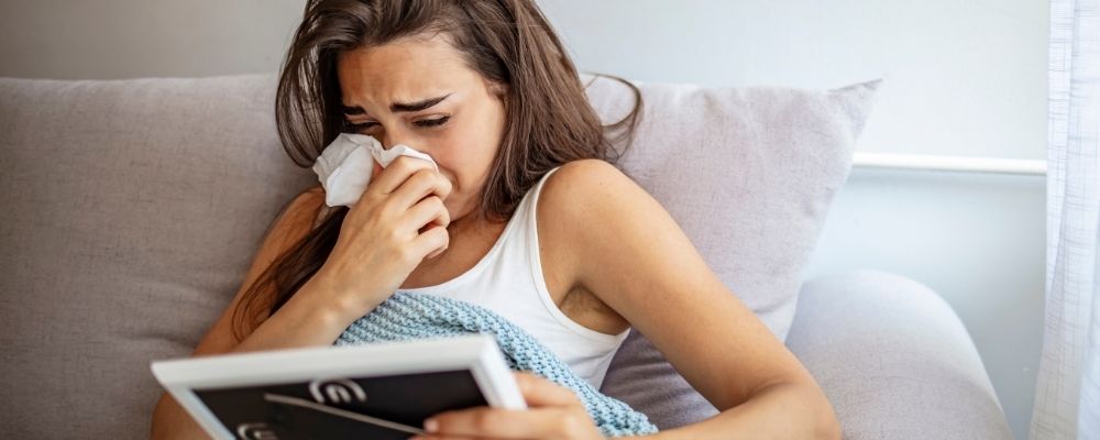 Vrouw die haar neus snuit met een tissue terwijl ze op bed zit met een tablet