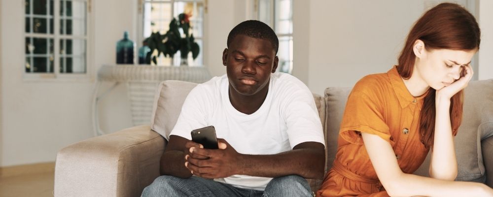 Man die op zijn telefoon bezig is en vrouw die teleurgesteld van hem wegkijkt door onzekerheid in een relatie
