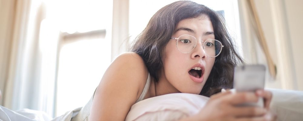 Vrouw die verrast naar haar telefoon kijkt terwijl ze op bed ligt
