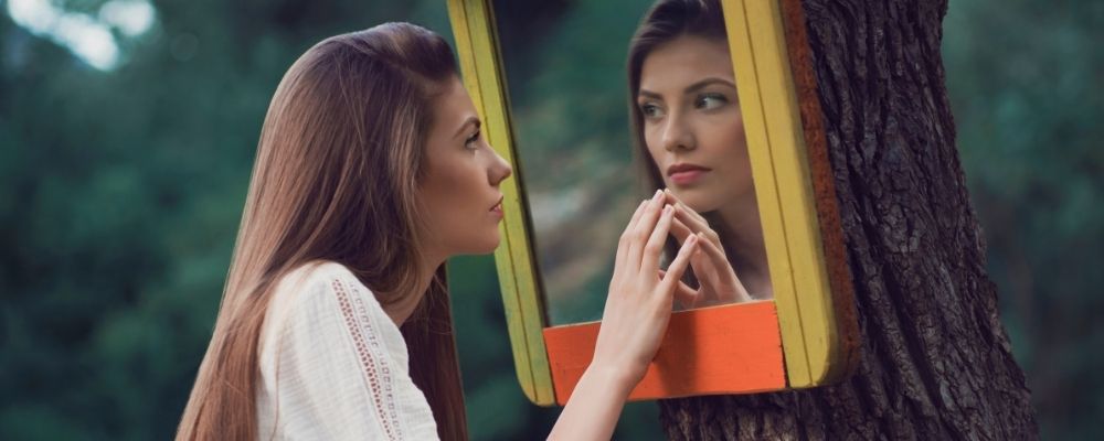 Vrouw die in het bos in een spiegel naar zichzelf kijkt en haar hand tegen de spiegel houdt