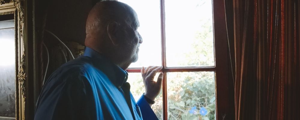Oudere man die eenzaam uit het raam kijkt en zijn hand tegen het raam houdt